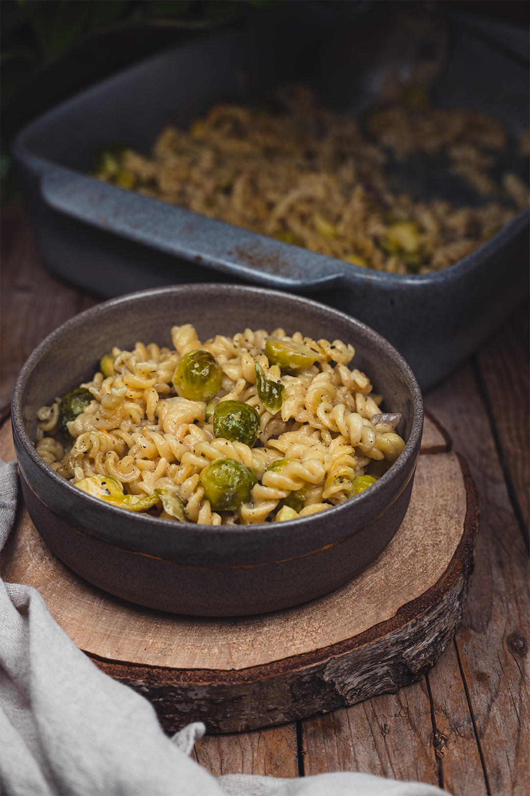 Auf einem hölzernen Untergrund liegt eine Holzscheibe, als Untersetzer für eine graue Keramik-Schüssel mit einem Goldenen Ring ringsherum. Darin befinden sich cremige Nudeln mit glänzendem Rosenkohl. Im Hintergrund ist eine graue Auflaufform zu erkennen, in der das Gericht im Ofen gegart wurde. Im vorderen Teil des Bildes liegt eine Leinenserviette. Die Stimmung des Bildes ist dunkel gemütlich.
