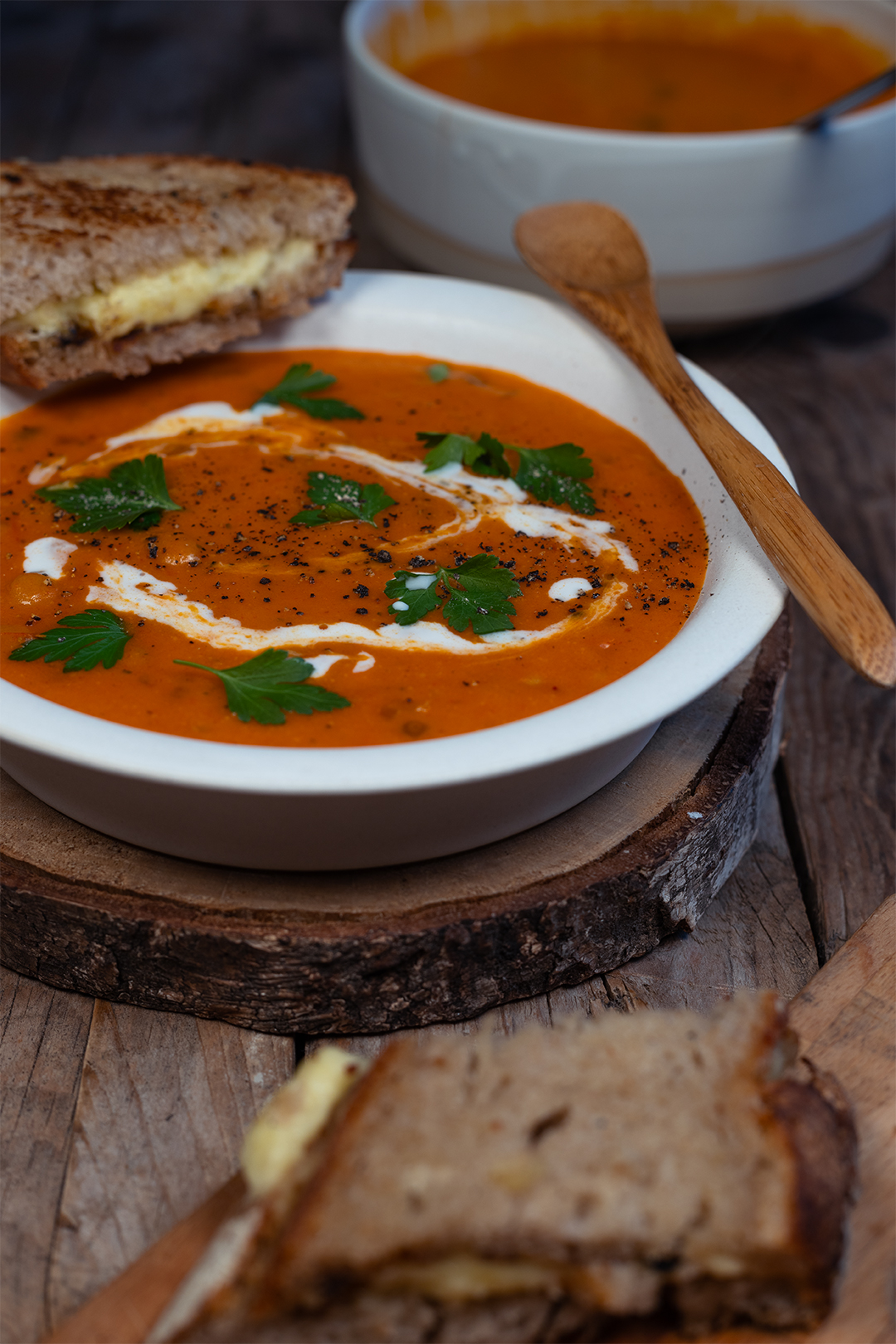 Auf einem hölzernen Untergrund liegt eine Holzscheibe, als Untersetzer für eine graue Keramik-Schüssel mit einem Goldenen Ring ringsherum. Darin befinden sich cremige Nudeln mit glänzendem Rosenkohl. Im Hintergrund ist eine graue Auflaufform zu erkennen, in der das Gericht im Ofen gegart wurde. Im vorderen Teil des Bildes liegt eine Leinenserviette. Die Stimmung des Bildes ist dunkel gemütlich.
