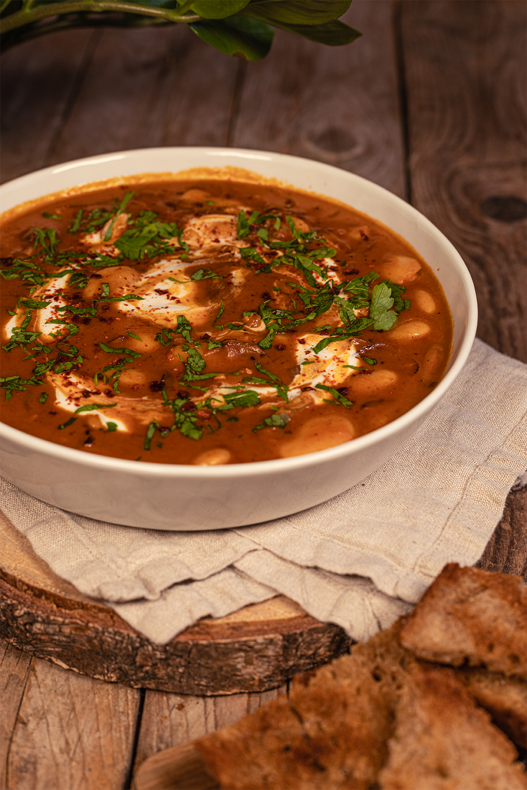 Auf einem hölzernen Untergrund steht eine Holzscheibe als Untergrund für eine Weiße Schüssel mit einer herrlichen Portion Tomaten-Kokos-Weiße-Bohnen Pfanne. nahe dabei liegen ein paar geröstete Brotscheiben. Das vegane Gericht ist mit pflanzlichem Joghurt, frischer Petersilie und ein paar Chiliflocken garniert. Unter der Schüssel liegt eine Leinenserviette. Die Stimmung des Bildes ist dunkel, gemütlich.