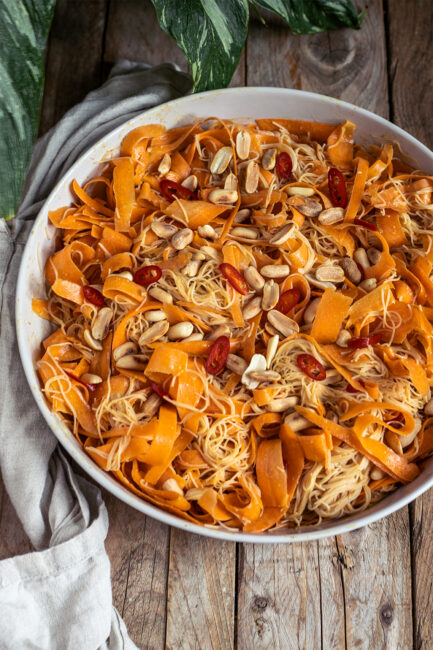 Veganer Möhren-Glasnudelsalat asiatisch: breite Karottenbänder und Glasnudeln mit Erdnussdressing, Toppings aus Erdnüssen und roter Chili, angerichtet in einer weißen Schale