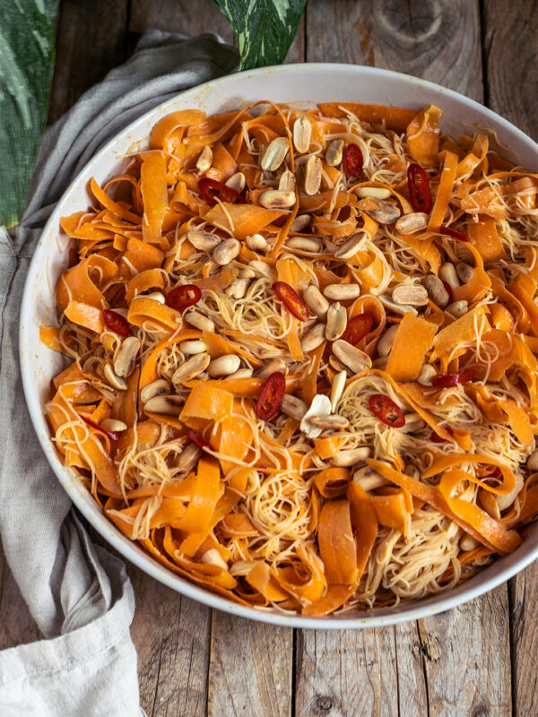 Veganer Möhren-Glasnudelsalat asiatisch: breite Karottenbänder und Glasnudeln mit Erdnussdressing, Toppings aus Erdnüssen und roter Chili, angerichtet in einer weißen Schale