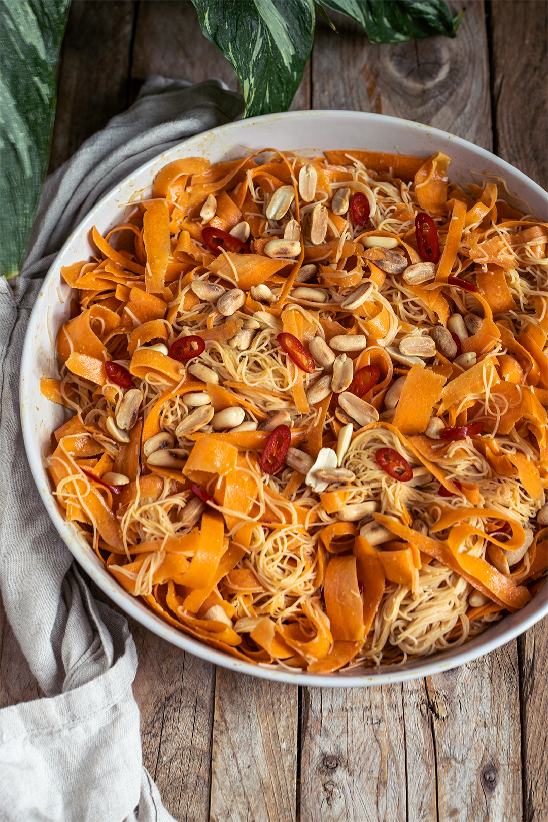 Veganer Möhren-Glasnudelsalat asiatisch: breite Karottenbänder und Glasnudeln mit Erdnussdressing, Toppings aus Erdnüssen und roter Chili, angerichtet in einer weißen Schale