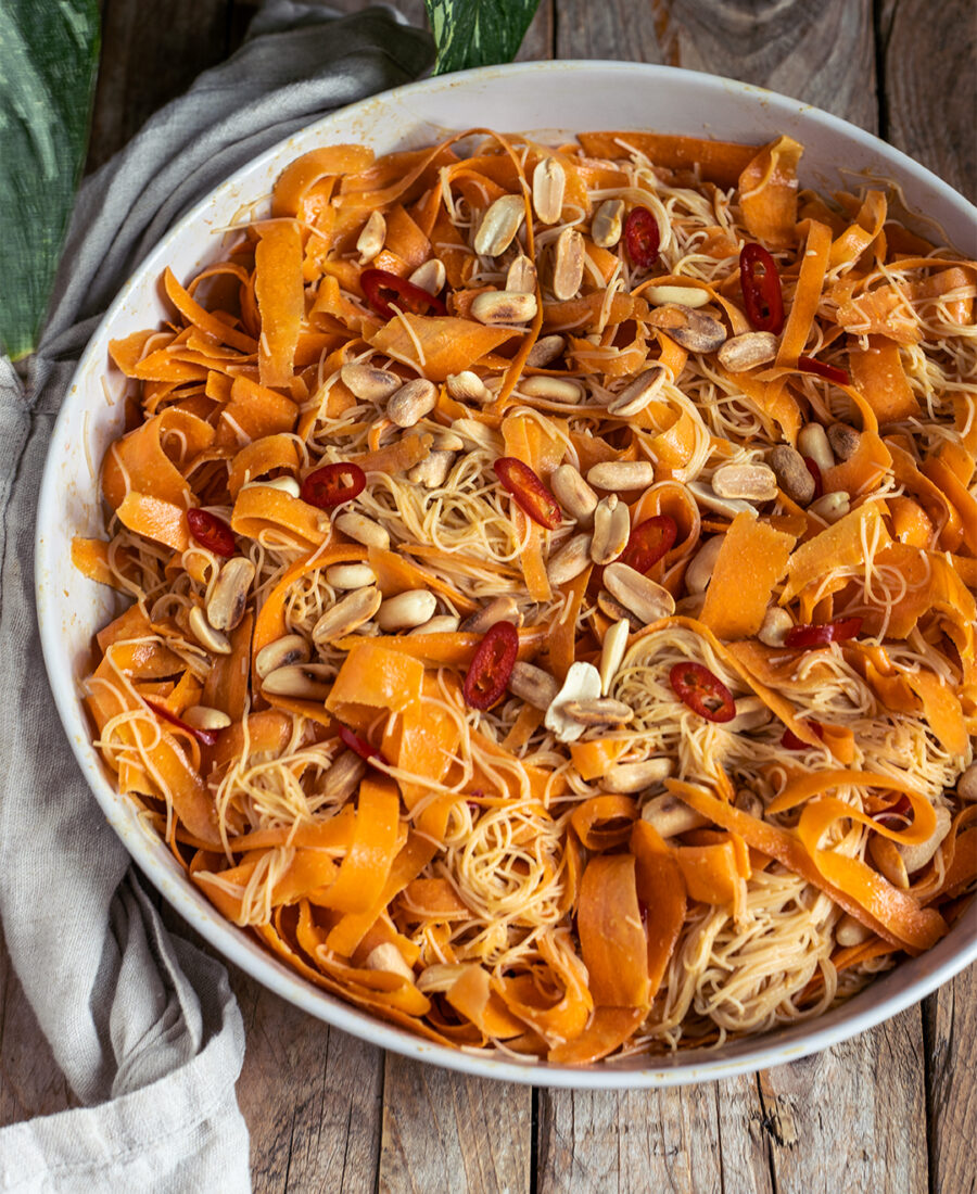 Veganer Möhren-Glasnudelsalat asiatisch: breite Karottenbänder und Glasnudeln mit Erdnussdressing, Toppings aus Erdnüssen und roter Chili, angerichtet in einer weißen Schale